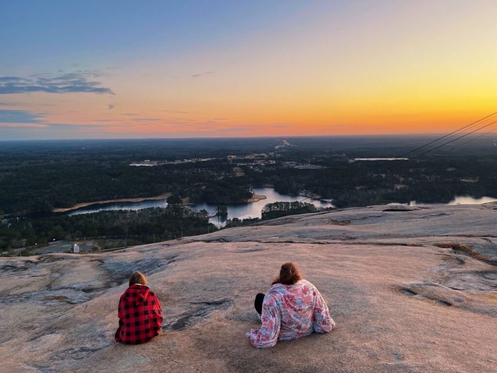 two girls bundled up on top of Stone Mountain overlooking a landscape of water and trees while the sun is rising