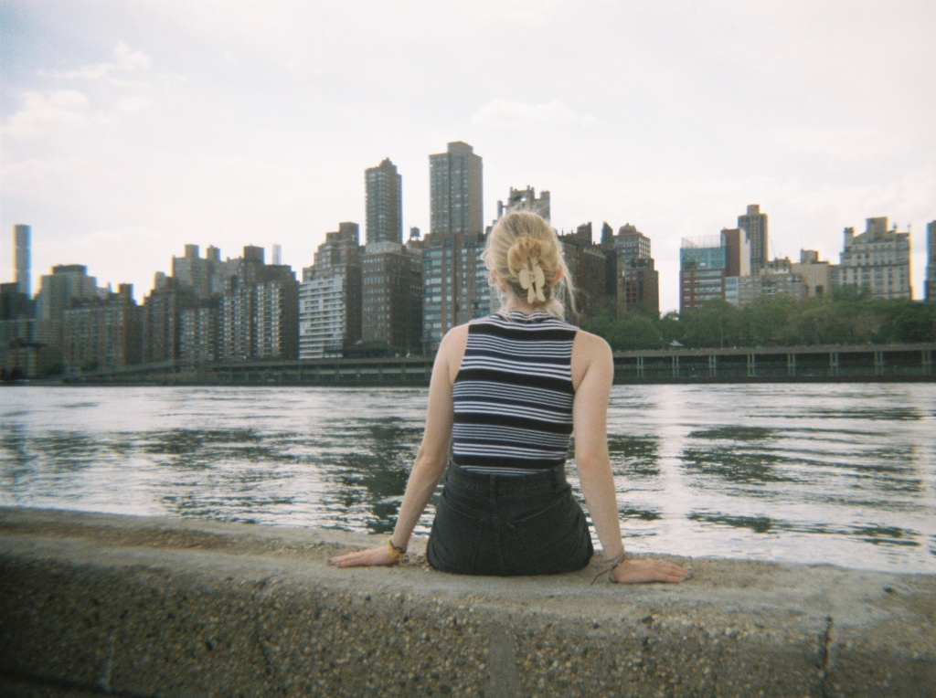 girl sitting on Roosevelt Island with the Manhattan skyline in the background 