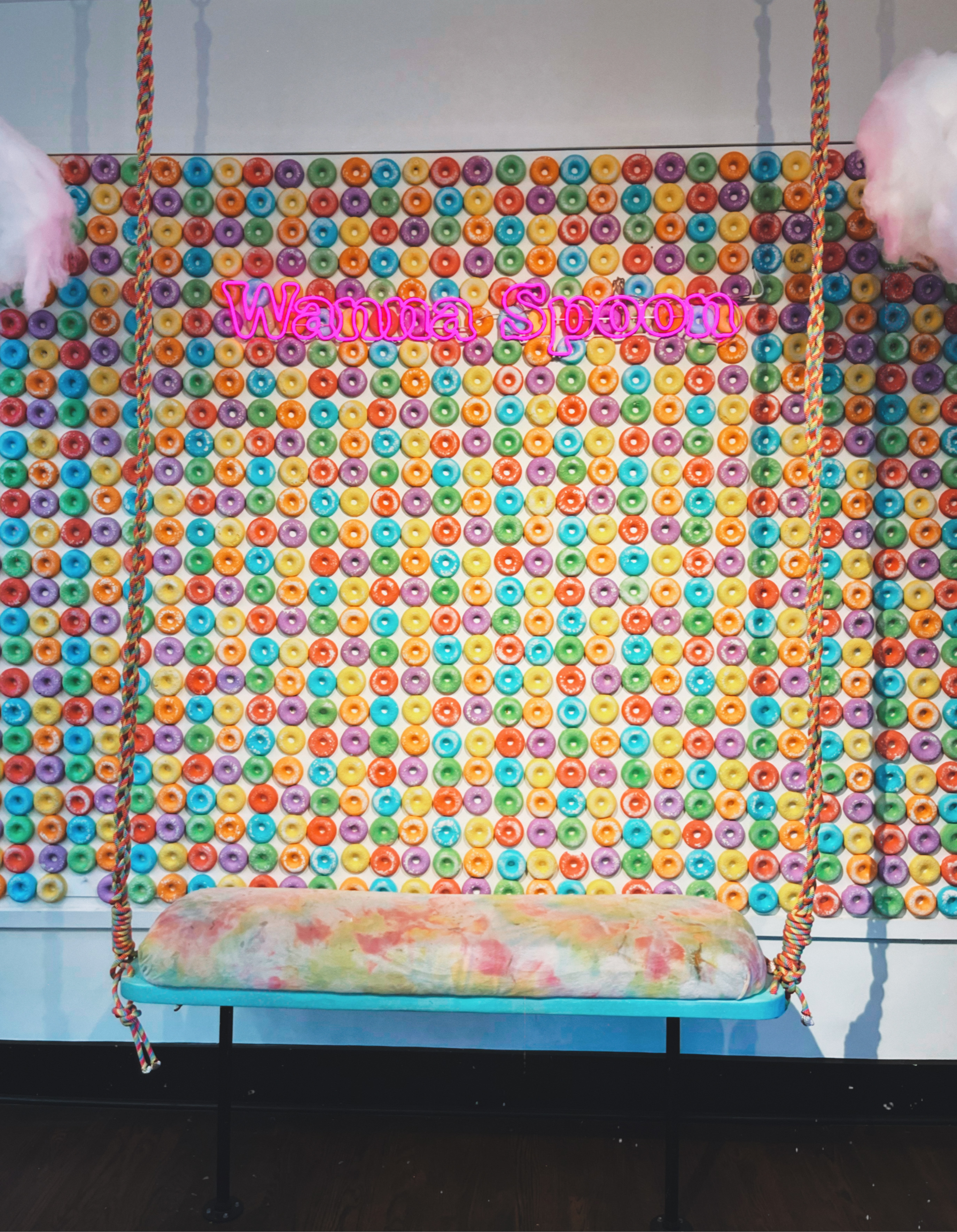 wall made of giant fruit loops with a neon sign that says wanna spoon, there is also a swing and fluffy clouds hanging from the ceiling 
