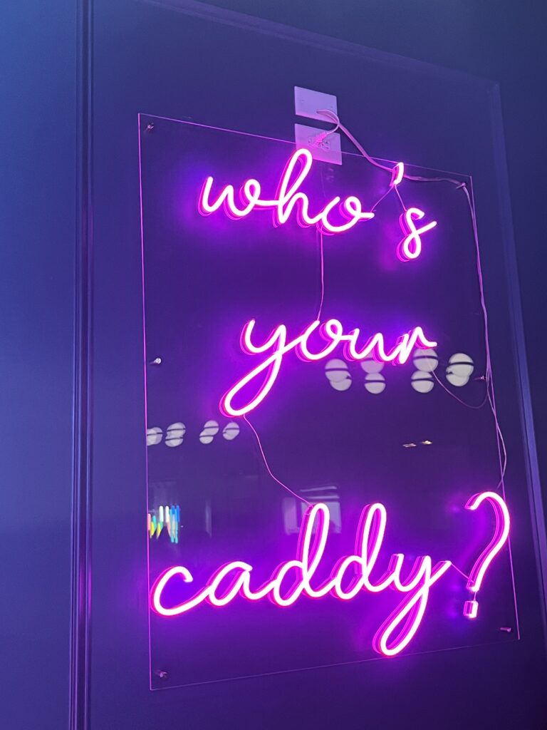 A neon sign that says "Who's Your Caddy" inside of Group Therapy" Pub & Playground