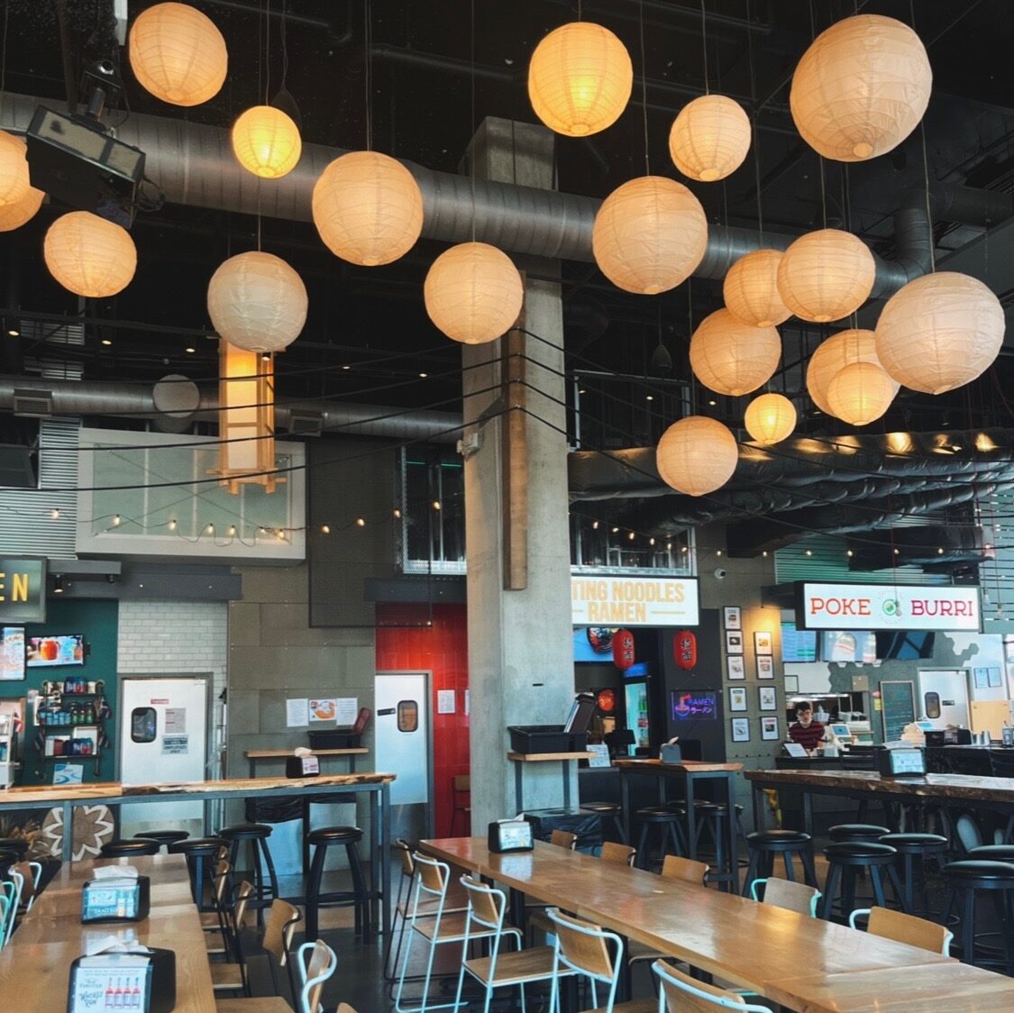 a picture of some of the communal tables and a few of the restaurants found in the Ph'east dining hall including Poke Burri. Above the tables are round lanterns. 