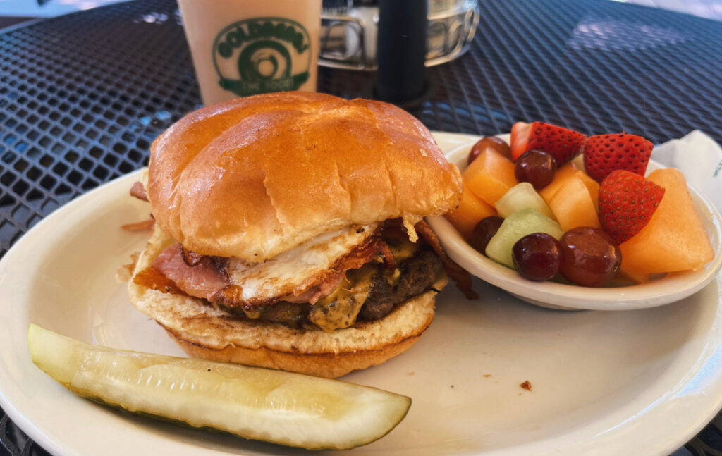 a Goldberg's breakfast sandwich with a hamburger patty, egg, cheese, and bacon with bowl of fruit on the side and a pickle. I also got a caramel latte which can be seen in the background.