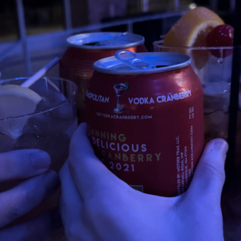 A picture of two canned alcohol drinks and two cocktails being clinked together in cheers